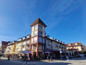 Ein mehrstöckiges Gebäude mit einem markanten Turm, Außenbestuhlung und blauem Himmel.