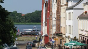 Une vue de rue avec des bâtiments, des voitures garées et une étendue d’eau en arrière-plan.