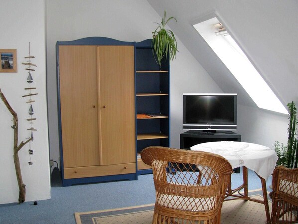Quarto com teto inclinado, guarda-roupa grande, televisão, cadeiras de vime e uma pequena mesa.