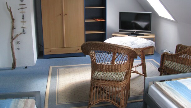 Une pièce avec un plafond en pente, une télévision, des fauteuils en osier, un lit et une armoire.