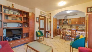 Uma sala de estar com sofá, uma poltrona verde, espaço de refeições com mesa e cadeiras, e cozinha com armários de madeira e micro-ondas.