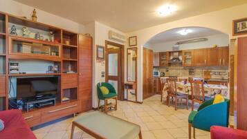 Un salon avec un canapé, un fauteuil vert, un coin repas avec une table et des chaises, et une cuisine avec des meubles en bois et un four à micro-ondes.