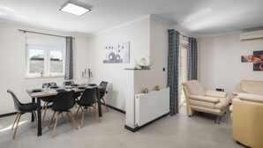 Moderni olohuone, jossa on ruokailutila. Tilaan kuuluu neljälle katettu ruokapöytä, beige sohva ja patteri.