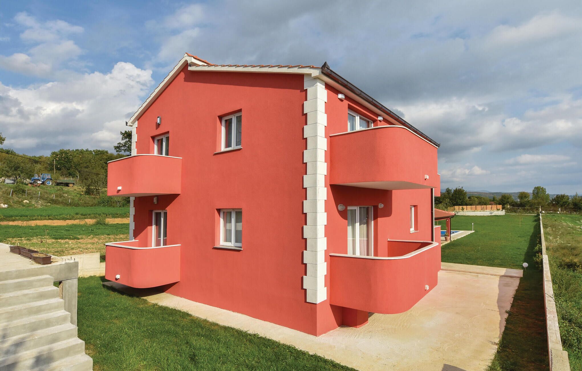 A red two-story house with a balcony on the second floor, situated in a rural area with green fields and a clear sky.