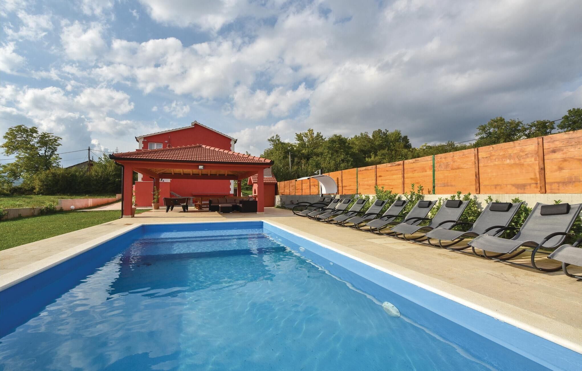 A swimming pool with lounge chairs, a covered seating area, and a red building in the background.