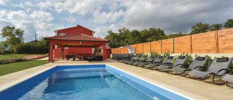 A swimming pool with lounge chairs, a covered seating area, and a red building in the background.