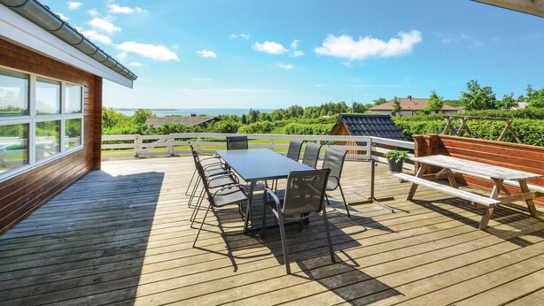 A wooden deck with a table and chairs, a bench, and a view of the sea and greenery. - Cozy home in Spøttrup with sauna (Spøttrup)