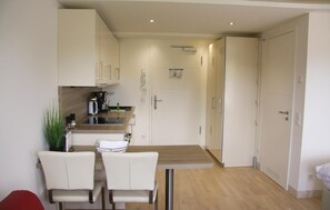 Una cocina compacta con armarios blancos, una mesa de comedor con sillas y una pequeña cafetera.