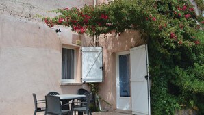 Une terrasse extérieure avec une table et des chaises, une porte blanche et une fenêtre avec un rideau.
