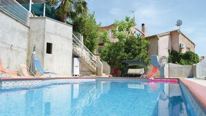 Une piscine dotée de chaises longues et d’un espace de repos couvert, le tout entouré de bâtiments et de verdure.