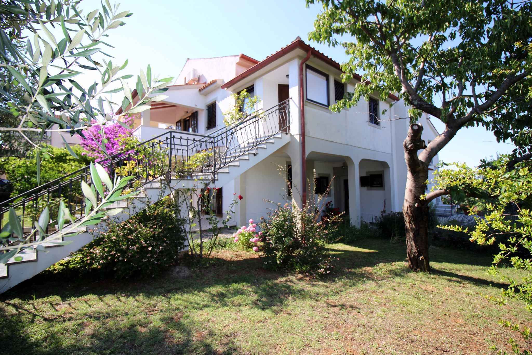 A two-story house with a staircase leading to the upper floor, surrounded by greenery and a tree.