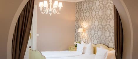 A double bed with white linens, a chandelier, a nightstand with a lamp, and a patterned wallpaper.