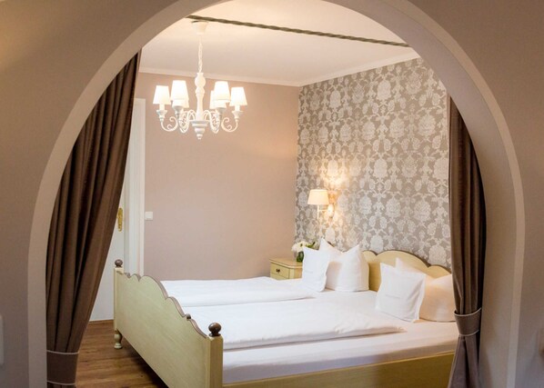 A double bed with white linens, a chandelier, a nightstand with a lamp, and a patterned wallpaper.