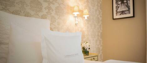 Una habitación de hotel con cama, dos almohadas blancas, una mesita de noche con un jarrón de flores y un cuadro en la pared.