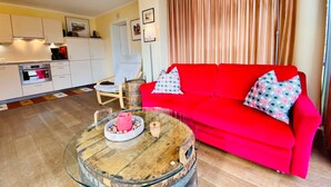 A living room with a red sofa, a wooden coffee table, a kitchen in the background, and a curtain.