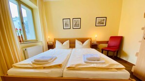 A double bed with white linens, a wooden headboard, a red chair, and two framed pictures on the wall.