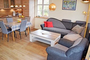 Ein modernes Wohnzimmer mit einem grauen Sofa, einem Esstisch mit Stühlen und einer Küche im Hintergrund.
