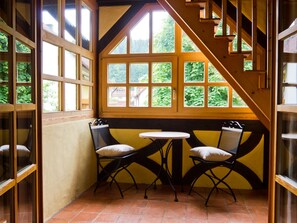 Habitación de esquina con ventana, dos sillas, una mesita y una escalera de madera.