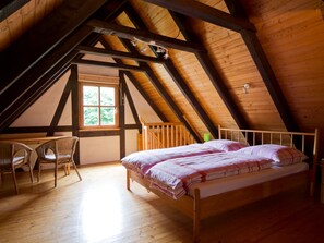 Ein Zimmer mit schrägem Dach, Holzbalken, einem Bett mit rosa Bettdecke, zwei Korbstühlen und einem Fenster mit Blick auf Grünflächen.