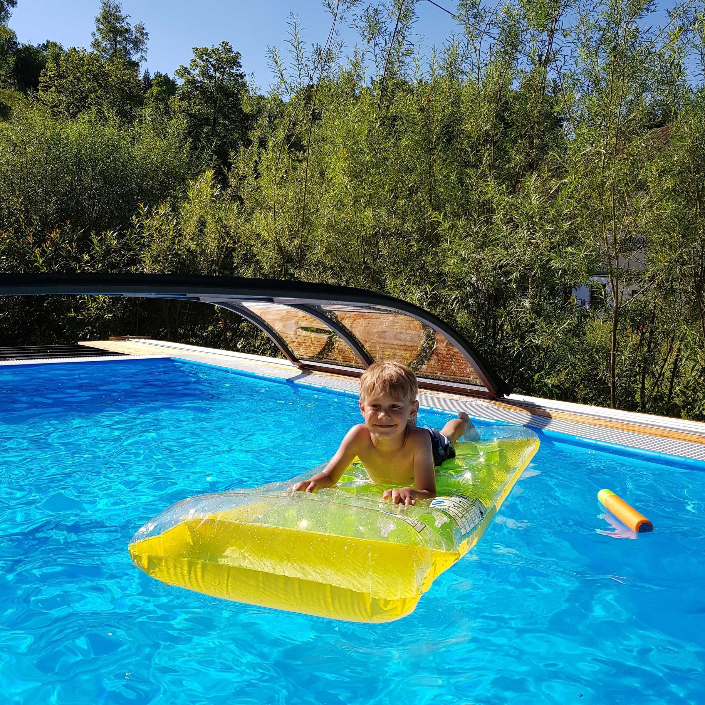 A child in a pool with a floating mat, surrounded by trees and a covered area.