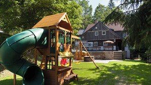 Un terrain de jeux avec un toboggan et des balançoires, entouré d’une pelouse et avec un bâtiment traditionnel en arrière-plan.