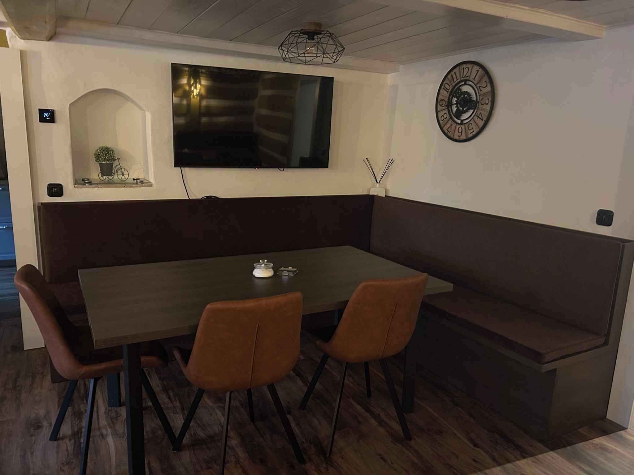 A dining area with a dark wooden table, two brown chairs, a bench, a wall-mounted clock, and a TV screen.