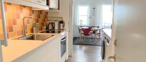 A compact kitchen with a sink, stove, and cabinets. A dining area with a table and chairs is visible through an open door.