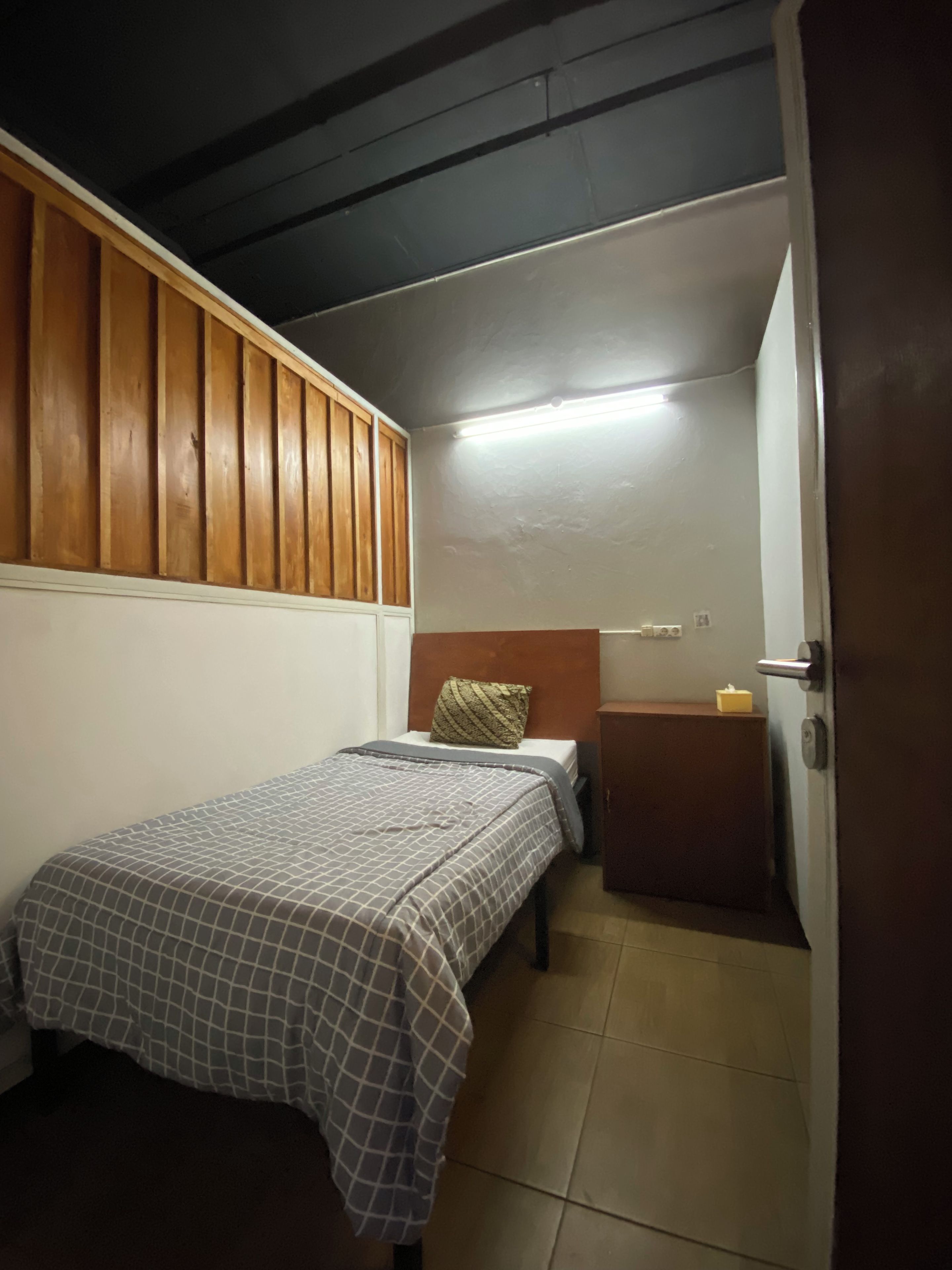 Ein kleines, gemütliches Zimmer mit einem Bett, einem Holz-Kopfstück, einem Nachttisch mit Lampe und einer Tür.
