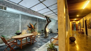 Um espaço amplo e interno, com teto alto, mesa e cadeiras de madeira, plantas em vasos e parede de pedra.