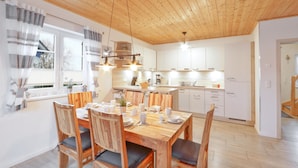Een moderne keuken met houten kastjes, een eethoek met een houten tafel en stoelen, en uitzicht op de omgeving door grote ramen.