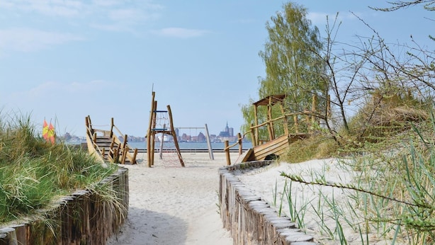 Een zandpad langs het strand dat leidt naar een houten constructie, met op de achtergrond een uitzicht op een stadsbeeld.