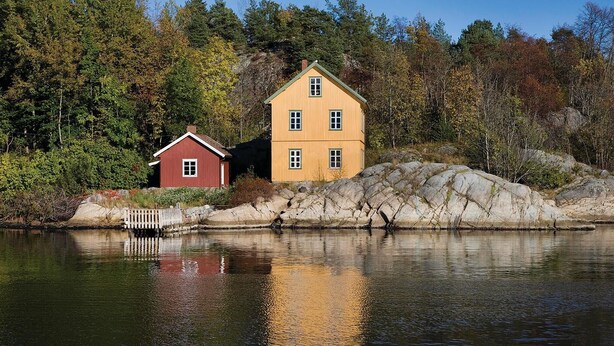 A two-story house with a red and yellow exterior, situated on a rocky shoreline with a body of water in the foreground.