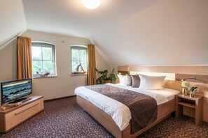 Une chambre avec un grand lit, un téléviseur posé sur une table de chevet en bois, deux fenêtres avec des rideaux et une plante en pot.