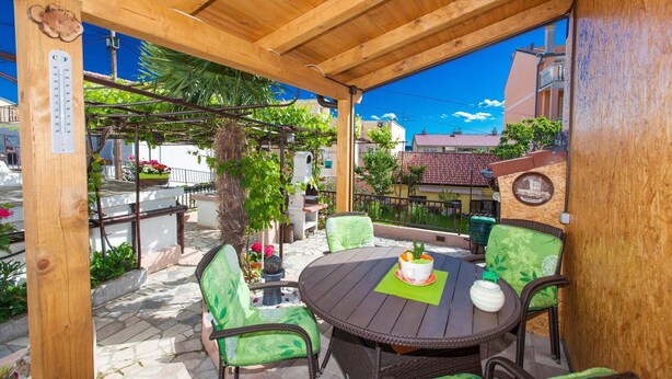An outdoor patio with a wooden pergola, green cushioned chairs, a round table, and a decorative plate on the wall.