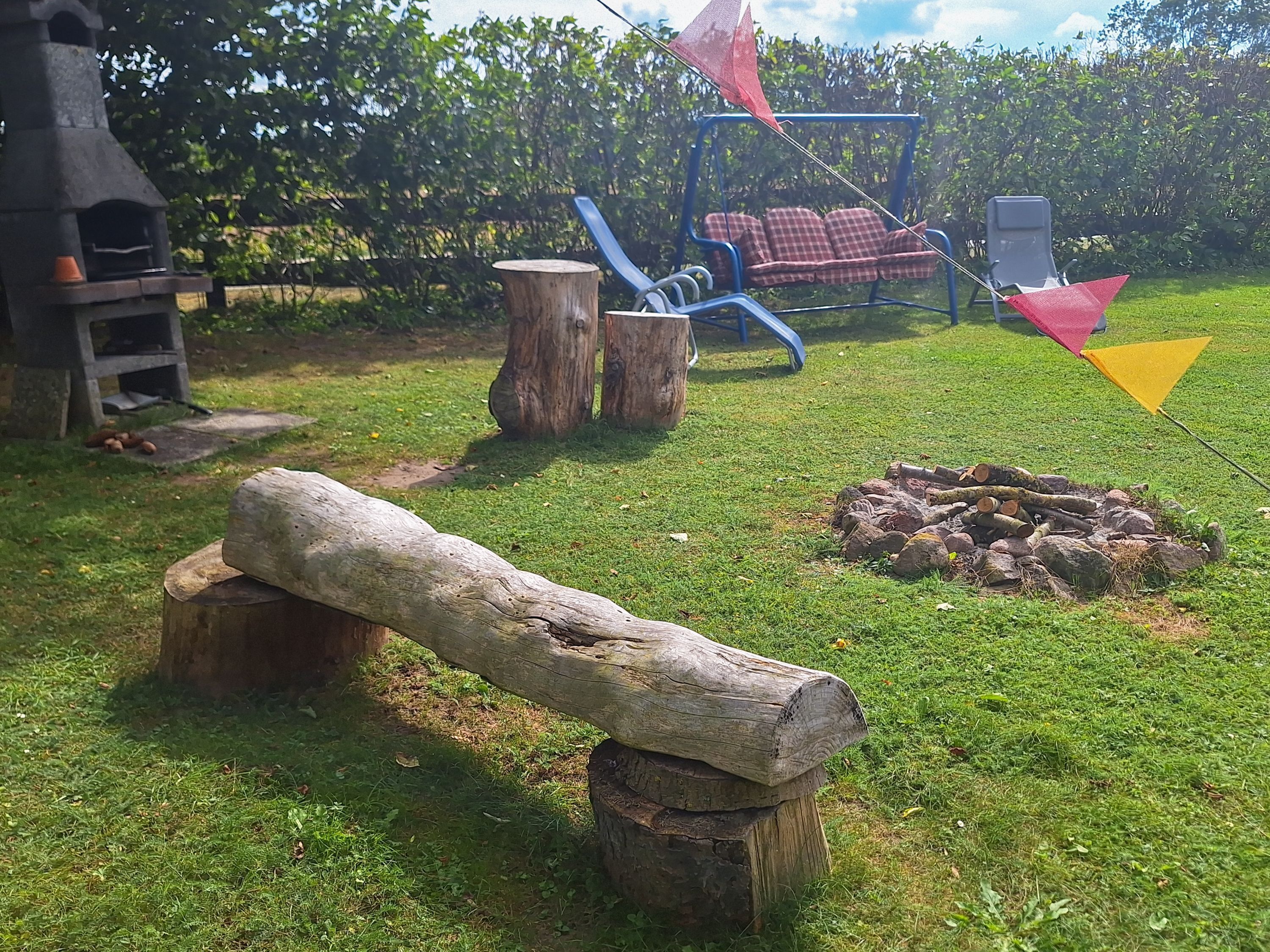 Eine ländliche Outdoor-Szenerie mit einem Holzbalken, einem Holzstoß, einer blauen Schaukel und einer rot-gelben Flagge.