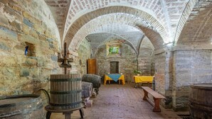 Un bâtiment médiéval en pierre, doté de voûtes en arc, de barils en bois et d’une table recouverte d’une nappe jaune.