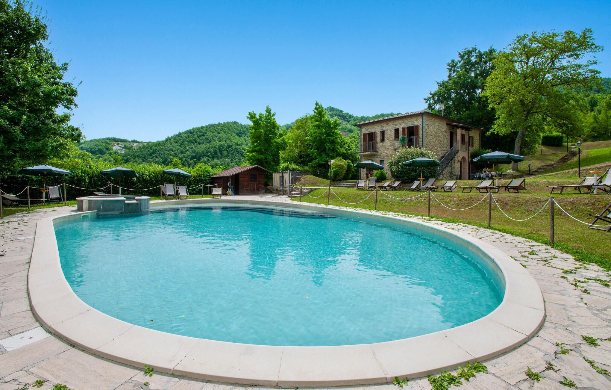 Une piscine entourée de verdure et d’un bâtiment en pierre, avec des chaises longues et des parasols à proximité.