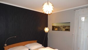 A bedroom with a wooden bed, white bedding, a wall-mounted lamp, and a beach scene painting. - * FeWo Hansel GM 69372 - FeWo Hansel (Graal-Müritz)