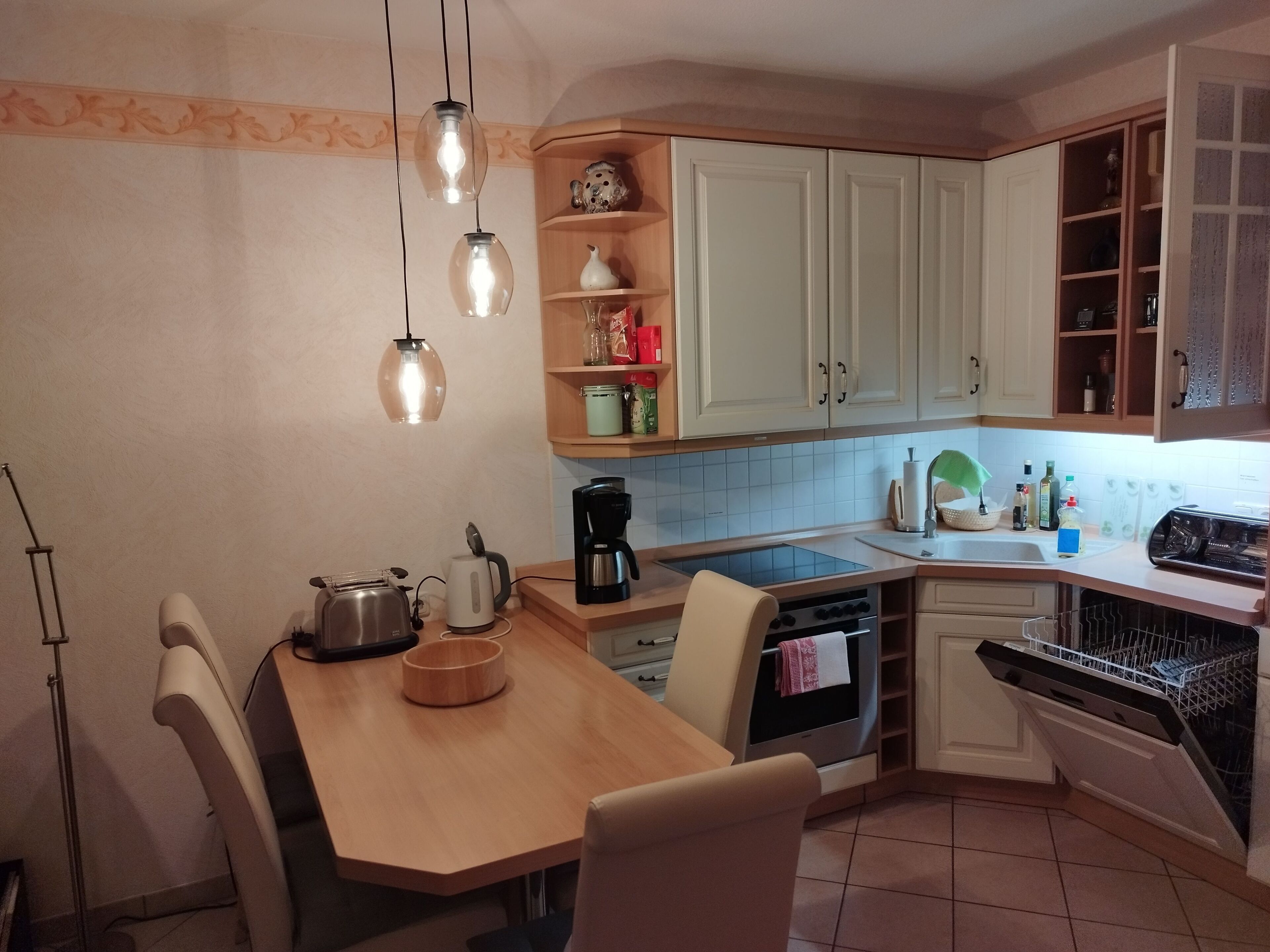 A compact kitchen with a wooden table, chairs, a coffee maker, a kettle, and a dishwasher.
