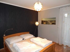 Une chambre à coucher avec un lit en bois, du linge de lit blanc, une lampe fixée au mur et un tableau sur le thème de la plage.