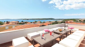 Une terrasse avec des meubles blancs, une table avec des boissons et une vue sur la mer et les bâtiments.