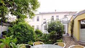 A patio area with a round glass table and wicker chairs, surrounded by greenery and a building in the background.