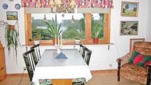 Ein Essbereich mit Tisch und Stühlen, ein Fenster mit Blick auf Grünflächen und ein Sessel mit gemustertem Kissen.