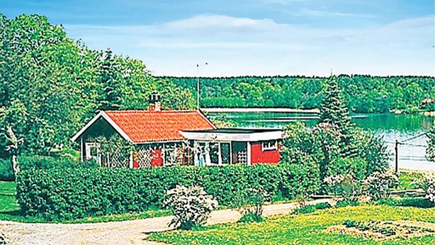Ein Haus mit rotem Dach und einer kleinen Veranda, umgeben von üppiger Vegetation und einem Gewässer im Hintergrund.