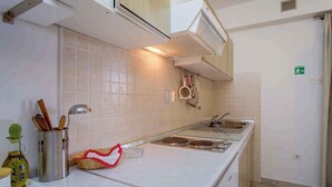 Cozinha com bancadas brancas, pia e fogão. Há um frasco de azeite, uma cesta e um recipiente de metal com utensílios.