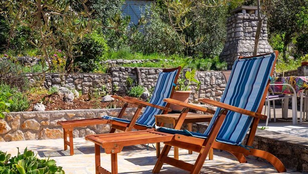Uma área de pátio com cadeiras de lounge de madeira e uma mesa, cercada por paredes de pedra e vegetação.