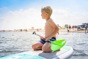 A person sitting on a paddleboard in a body of water with a beach and people in the background. - Holiday home with terrace near the beach (Damp)