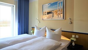 Une chambre d’hôtel avec un grand lit, une fenêtre avec des rideaux bleus, un tableau représentant une plage accroché au mur et une table de chevet sur laquelle est posé un vase de fleurs.