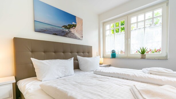 A bedroom with a large bed, a headboard, a window with a view of greenery, and a painting on the wall.