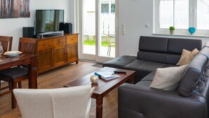 A living room with a sofa, a wooden coffee table, a dining table with chairs, a television on a wooden cabinet, and a window with a plant and a heart-shaped decoration.